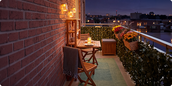 Mały balkon z drewnianymi krzesłami i stołem, oświetlony lampionami i świecami, udekorowany kwiatami w doniczkach i zielenią, z widokiem na panoramę miasta nocą.