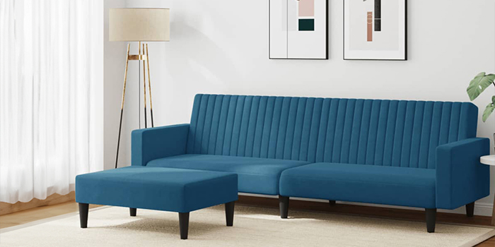 Nowoczesna niebieska sofa narożna z aksamitu z pionowym pikowaniem kanałowym i pasującym podnóżkiem. Sofa ma czarne drewniane nogi i stoi w jasnym salonie z oprawionymi obrazami na ścianie oraz lampą podłogową w rogu.